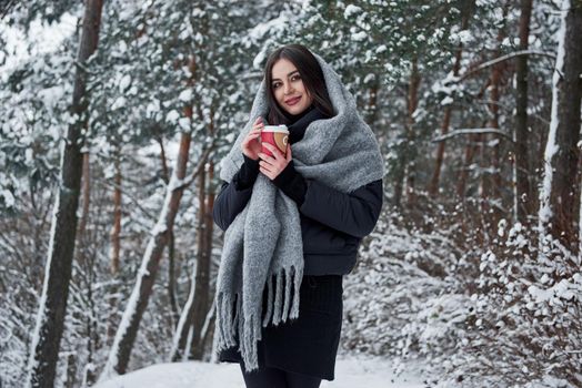 Looking into the camera. Girl in warm clothes with cup of coffee have a walk in the winter forest.