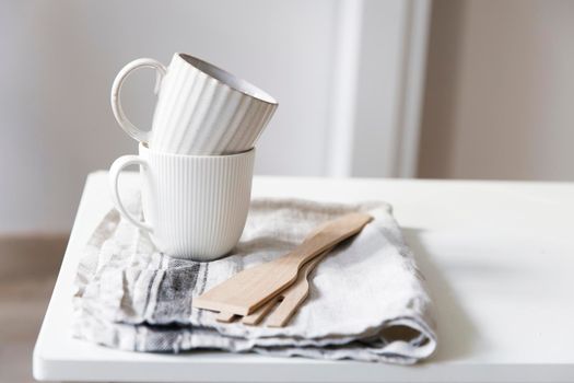 Two white mugs, a kitchen towel, a napkin and wooden frying utensils on the table. Defocus.