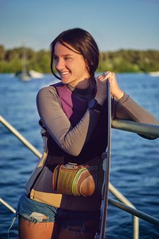 Alluring brunette girl in a gray turtleneck, special sports vest and black watch is looking away and smiling while posing with her wakeboard and standing on a pier of the riverside. Sport and recreation concept. Close-up shot.