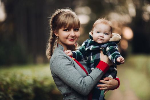 Professional portrait of attractive young woman with fair hair in braid embracing her little baby girl in plaid warm overall while standing in beautiful bright tree with red leaves. They are smiling at camera surrounded by vivid foliage in autumnal park.