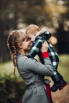 Professional portrait of attractive young woman with fair hair in braid embracing her little baby girl in plaid warm overall while standing in beautiful bright tree with red leaves. They are smiling at camera surrounded by vivid foliage in autumnal park.