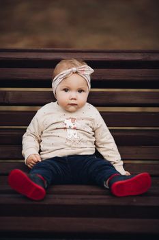 Portrait of lovely little baby girl wearing cute jersey with bunny on it and beige patterned bow headband. She is smiling at camera while parents holding her arms. Weekend in the park in autumn. Baby doing her first steps.