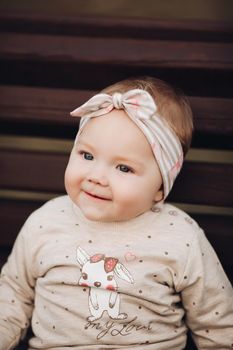 Portrait of lovely little baby girl wearing cute jersey with bunny on it and beige patterned bow headband. She is smiling at camera while parents holding her arms. Weekend in the park in autumn. Baby doing her first steps.