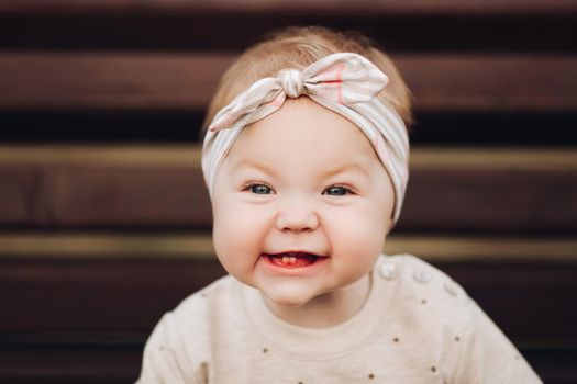 Portrait of lovely little baby girl wearing cute jersey with bunny on it and beige patterned bow headband. She is smiling at camera while parents holding her arms. Weekend in the park in autumn. Baby doing her first steps.