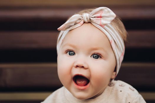 Portrait of lovely little baby girl wearing cute jersey with bunny on it and beige patterned bow headband. She is smiling at camera while parents holding her arms. Weekend in the park in autumn. Baby doing her first steps.