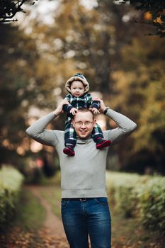 Professional portrait of attractive young man embracing his little baby girl in plaid warm overall while standing in beautiful bright tree with red leaves. They are smiling at camera surrounded by vivid foliage in autumnal park.