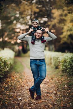 Professional portrait of attractive young man embracing his little baby girl in plaid warm overall while standing in beautiful bright tree with red leaves. They are smiling at camera surrounded by vivid foliage in autumnal park.