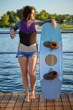 Alluring brunette female in a gray turtleneck, special sports vest and blue denim shorts is looking away while posing with her wakeboard and standing on a pier of the riverside. Sport and recreation concept. Full length shot.