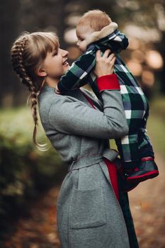 Professional portrait of attractive young woman with fair hair in braid embracing her little baby girl in plaid warm overall while standing in beautiful bright tree with red leaves. They are smiling at camera surrounded by vivid foliage in autumnal park.