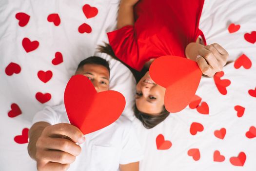 Top view of young attractive couple in love lying on the bed and showing red paper hearts. Happy valentine's day concept. High quality photo