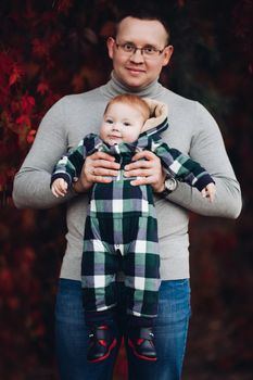 Professional portrait of attractive young man embracing his little baby girl in plaid warm overall while standing in beautiful bright tree with red leaves. They are smiling at camera surrounded by vivid foliage in autumnal park.