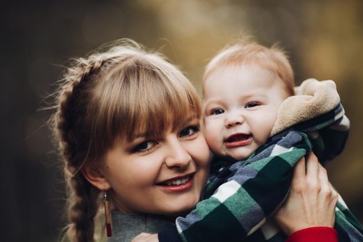 Professional portrait of attractive young woman with fair hair in braid embracing her little baby girl in plaid warm overall while standing in beautiful bright tree with red leaves. They are smiling at camera surrounded by vivid foliage in autumnal park.