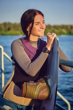 Alluring brunette woman in a gray turtleneck, special sports vest and black watch is looking away and smiling while posing with her wakeboard and standing on a pier of the riverside. Sport and recreation concept. Close-up shot.