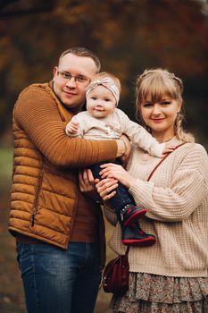 Portrait of attractive young mother and handsome smiling father wearing glasses holding their beautiful lovely baby girl on hands standing against green hedge in autumnal park. They are smiling and looking at camera.