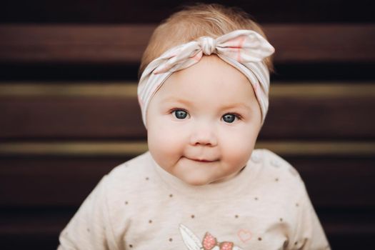 Portrait of lovely little baby girl wearing cute jersey with bunny on it and beige patterned bow headband. She is smiling at camera while parents holding her arms. Weekend in the park in autumn. Baby doing her first steps.