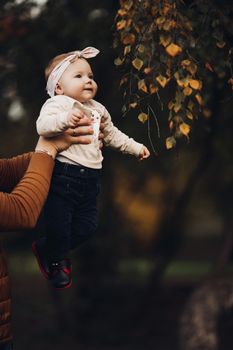 Portrait of lovely little baby girl wearing cute jersey with bunny on it and beige patterned bow headband. She is smiling at camera while parents holding her arms. Weekend in the park in autumn. Baby doing her first steps.