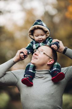 Professional portrait of attractive young man embracing his little baby girl in plaid warm overall while standing in beautiful bright tree with red leaves. They are smiling at camera surrounded by vivid foliage in autumnal park.