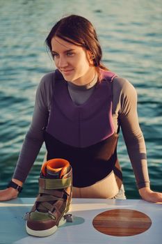 Attractive brunette female in a gray turtleneck and special sports vest is going to go down into the water while posing with her wakeboard on a pier of the coastal zone. Sport and recreation concept. Close-up shot.