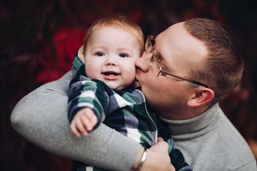 Professional portrait of attractive young man embracing his little baby girl in plaid warm overall while standing in beautiful bright tree with red leaves. They are smiling at camera surrounded by vivid foliage in autumnal park.