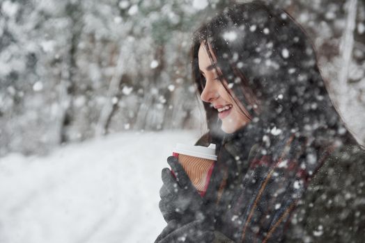 Falling snow in the woods. Girl in warm clothes with cup of coffee have a walk in the winter forest.
