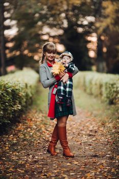 Professional portrait of attractive young woman with fair hair in braid embracing her little baby girl in plaid warm overall while standing in beautiful bright tree with red leaves. They are smiling at camera surrounded by vivid foliage in autumnal park.