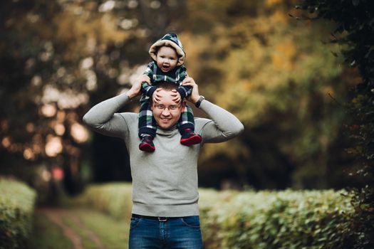 Professional portrait of attractive young man embracing his little baby girl in plaid warm overall while standing in beautiful bright tree with red leaves. They are smiling at camera surrounded by vivid foliage in autumnal park.