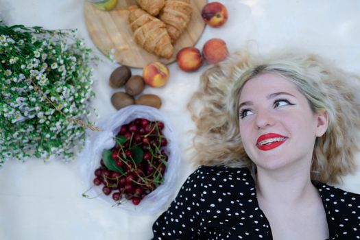 sweet blonde girl on picnic lying on white blanket in park. fresh summer refreshing drink and snacks. fruits and flowers.