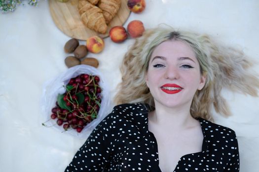 sweet blonde girl on picnic lying on white blanket in park. fresh summer refreshing drink and snacks. fruits and flowers.