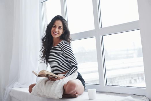 Smiling for the portrait. Girl reading book while sitting on the windowsill. Having free day.