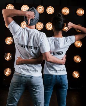 Gestures with thumbs up and hugs. Back view of couple in shirts with their names on it standing next to studio lights.