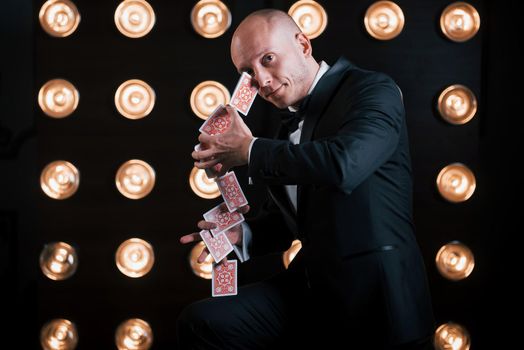 These cards is like he's second nature. Magician in black suit standing in the room with special lighting at backstage.
