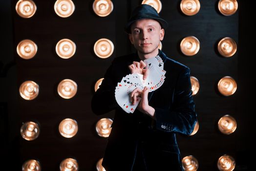 Conception of illusionist. Magician in black suit and with playing cards standing in the room with special lighting at backstage.