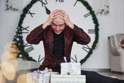 Too many presents. Man with big clock behind him sitting with New year gift boxes in holiday clothings.