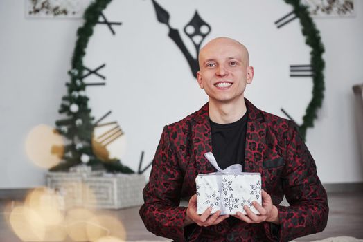 Smiling with present in hands. Man with big clock behind him sitting with New year gift boxes in holiday clothings.