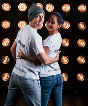 Turned to the camera. Back view of couple in shirts with their names on it standing next to studio lights.