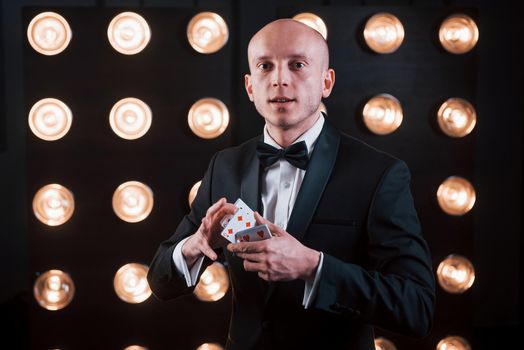 It's time to see some awesome trick. Magician in black suit standing in the room with special lighting at backstage.