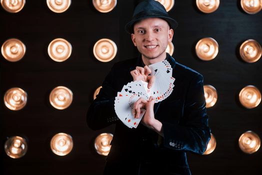 Your turn to pick. Magician in black suit and with playing cards standing in the room with special lighting at backstage.