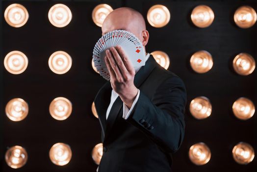 Focused photo on playing cards. Magician in black suit standing in the room with special lighting at backstage.