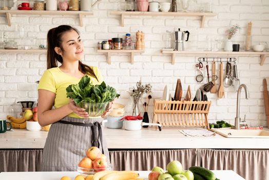 Preparing healthy foods. Healthy eating and dieting. Young caucasian brunette woman holding a bowl of fresh spinach in the kitchen