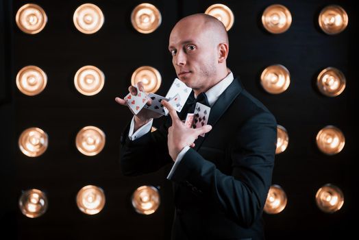 Hard professional tricks. Magician in black suit and with playing cards standing in the room with special lighting at backstage.