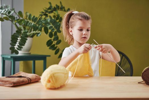 Kid is knitting at home. Cute little girl sitting near the wooden table is learning some new stuff.