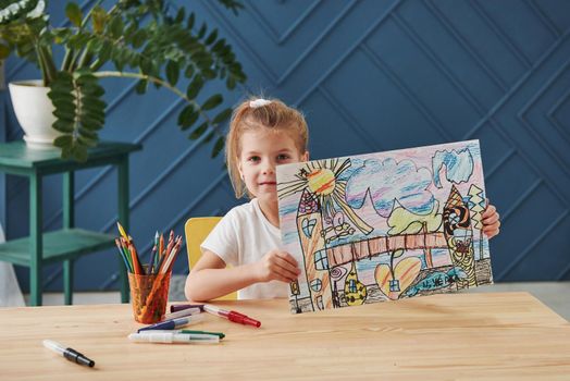 Nice interior in the room. Cute little girl in art school shows her first paintings drawn by pencils and markers.