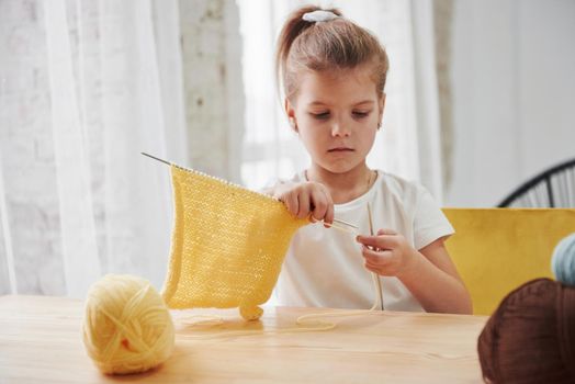 She's good at it. Kid is knitting at home. Cute little girl sitting near the wooden table is learning some new stuff.