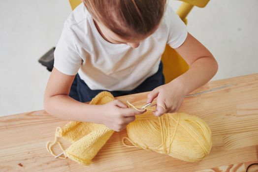 It will be such a nice cap. Kid is knitting at home. Cute little girl sitting near the wooden table is learning some new stuff.