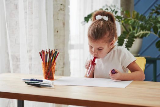 Holding marker in mouth. Cute little girl in art school draws her first paintings.