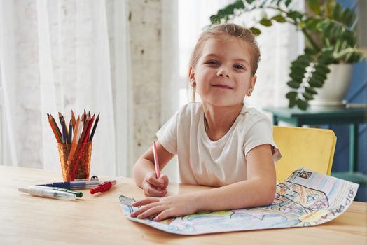 Looking into the camera with satisfied look. Cute little girl in art school draws her first paintings by pencils and markers.