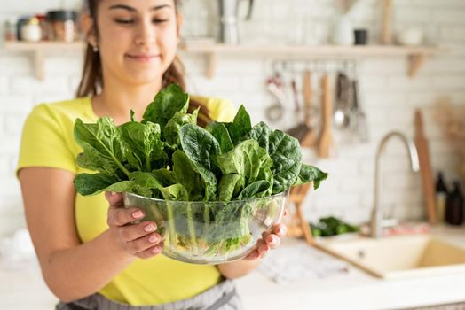 Preparing healthy foods. Healthy eating and dieting. Young caucasian brunette woman holding a bowl of fresh spinach in the kitchen