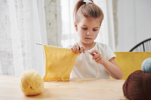 Craft and hobby for a child. Kid is knitting at home. Cute little girl sitting near the wooden table is learning some new stuff.