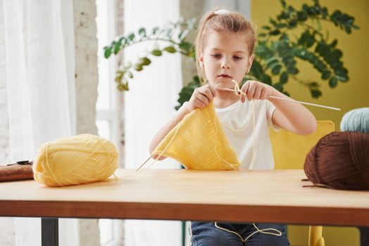 Looks like someone will have new cap. Kid is knitting at home. Cute little girl sitting near the wooden table is learning some new stuff.
