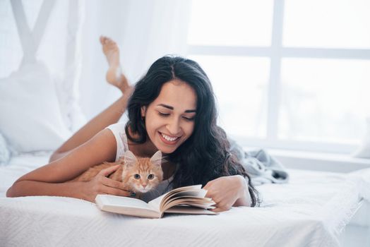 Home calm. Attractive blonde resting on the white bed with her cute kitten.